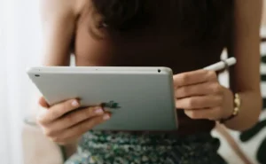 Uma mulher de blusa marrom segura um tablet e uma caneta "stylus", como se estivesse trabalhando em alguma ferramenta de marketing digital, seja escrevendo algo, ou trabalhando no design de alguma arte.