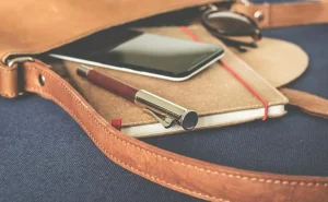 Uma bolsa é exibida aberta. e nota-se que de dentro dela estão saindo um pequeno caderno para anotações e uma caneta, além de um smartphone e um óculos escuro.
