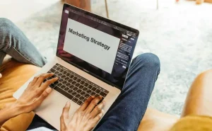 Uma pessoa com notebook no colo, com o texto "Marketing Strategy" (Estratégias de Marketing, em português) evidente na tela.