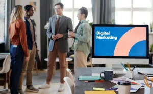 Quatro pessoas, vestidas como executivos, conversam ao fundo; enquanto em primeiro plano está em destaque um monitor com um slide aberto em tela cheia com o título "Digital Marketing".