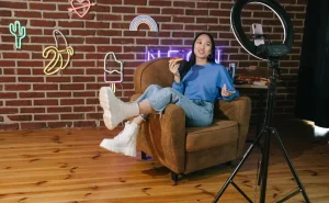 Uma influenciadora digital posa para spotlight em sua frente com um smartphone - provavelmente fazendo um conteúdo para suas redes sociais. No cenário temos uma parede de tijolos e emojis feitos de neon.