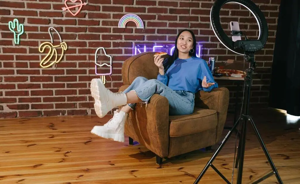 Uma influenciadora digital posa para spotlight em sua frente com um smartphone - provavelmente fazendo um conteúdo para suas redes sociais. No cenário temos uma parede de tijolos e emojis feitos de neon.
